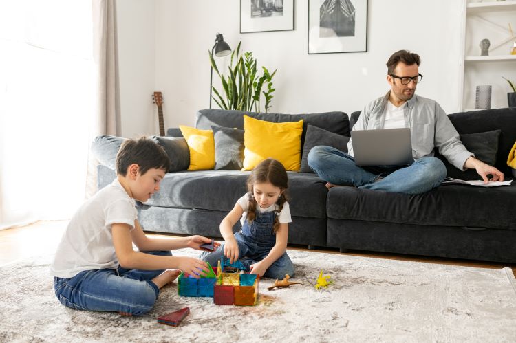 role models Father using laptop for freelance work sitting on the sofa at home while his two kids playing peacefully together on the floor. Busy single parent is babysitting and working on the distance
